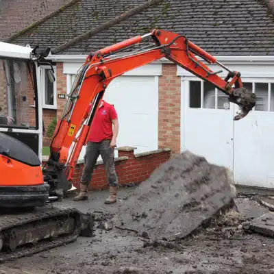 Construction worker and excavator removing old driveway surface outside a residential home for renovation