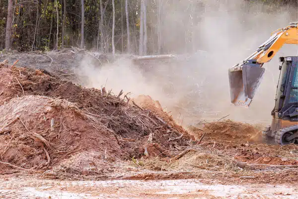 Excavator moves soil and uprooted trees, performing land clearing services on dusty wooded construction site