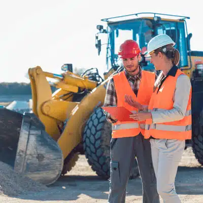 Engineers in safety vests discuss plans near loader, showcasing commercial excavation services managing large projects